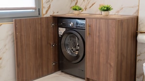 A compact, modern laundry room with a built-in washer and dryer, wooden cabinetry, marble flooring, and a small potted plant on the counter. - Lemair Luxury Aprtmnts Kozani Deluxe Junior Suite (Kozani)