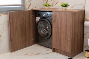 A compact, modern laundry room with a built-in washer and dryer, wooden cabinetry, marble flooring, and a small potted plant on the counter. - Lemair Luxury Aprtmnts Kozani Deluxe Junior Suite (Kozani)