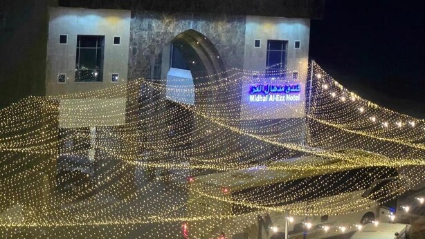 Um edifício com a placa 'Midhal Al-Ezz Hotel' e uma área de estacionamento com veículos.