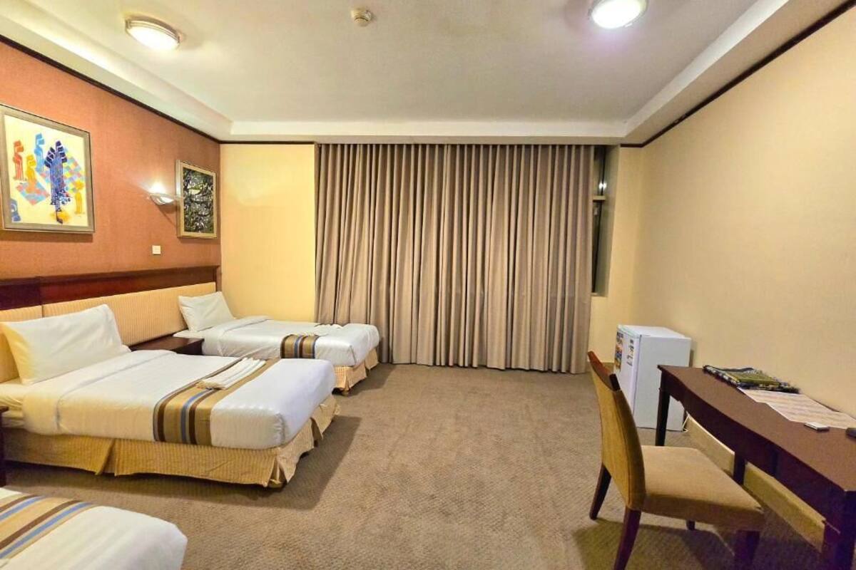 Hotellihuone, jossa on kolme sänkyä, työpöytä, tuoli, jääkaappi ja suuri ikkuna, jossa on verhot.