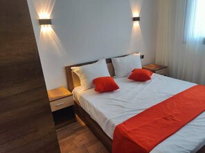 Ein Schlafzimmer mit hölzernem Kopfteil, zwei Nachttischen, einem Bett mit weißer Bettwäsche und roten Akzenten sowie Wandleuchten.