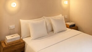 Ein ordentlich bezogenes Bett mit weißer Bettwäsche und Kissen, ein Nachttisch aus Holz mit Büchern sowie wandmontierte Leuchten.