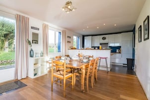 Una cocina moderna con armarios blancos, un comedor con mobiliario de madera y vistas al exterior con vegetación.