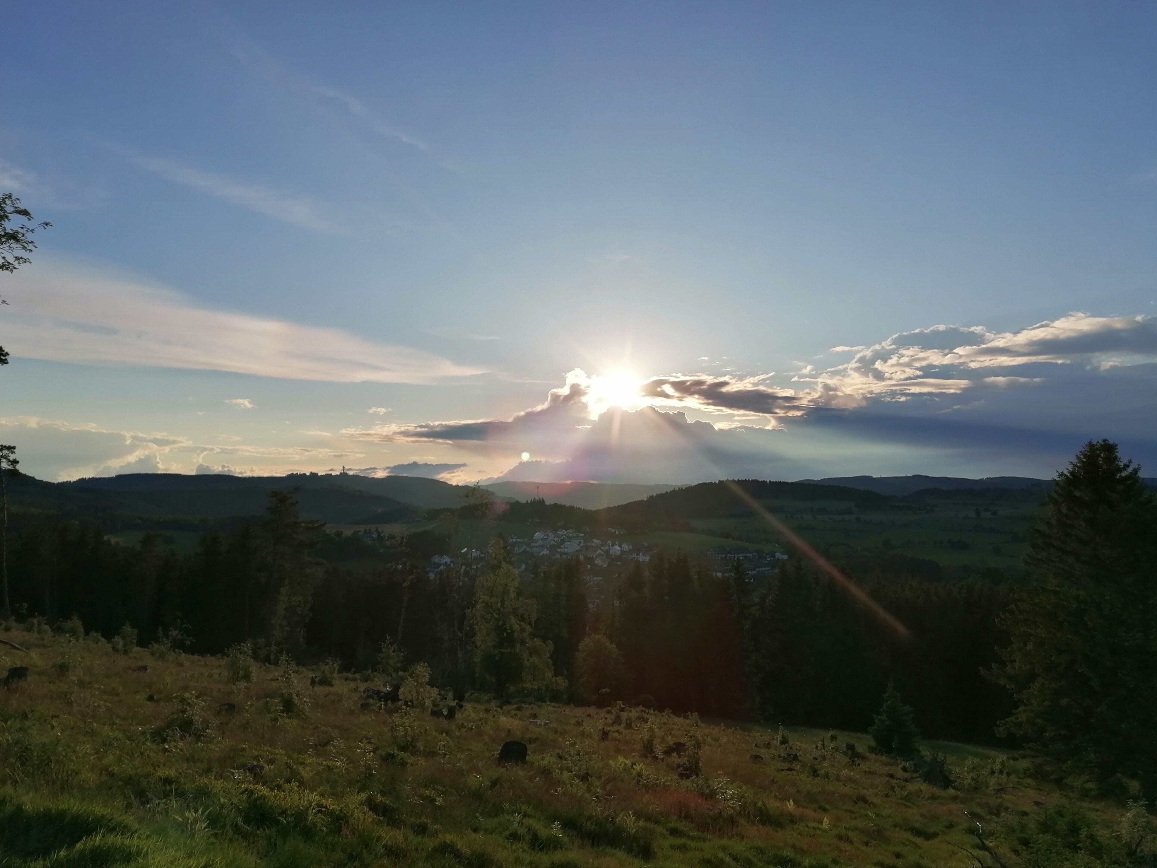 A landscape with a sun setting behind hills, a clear sky, and a forested area.