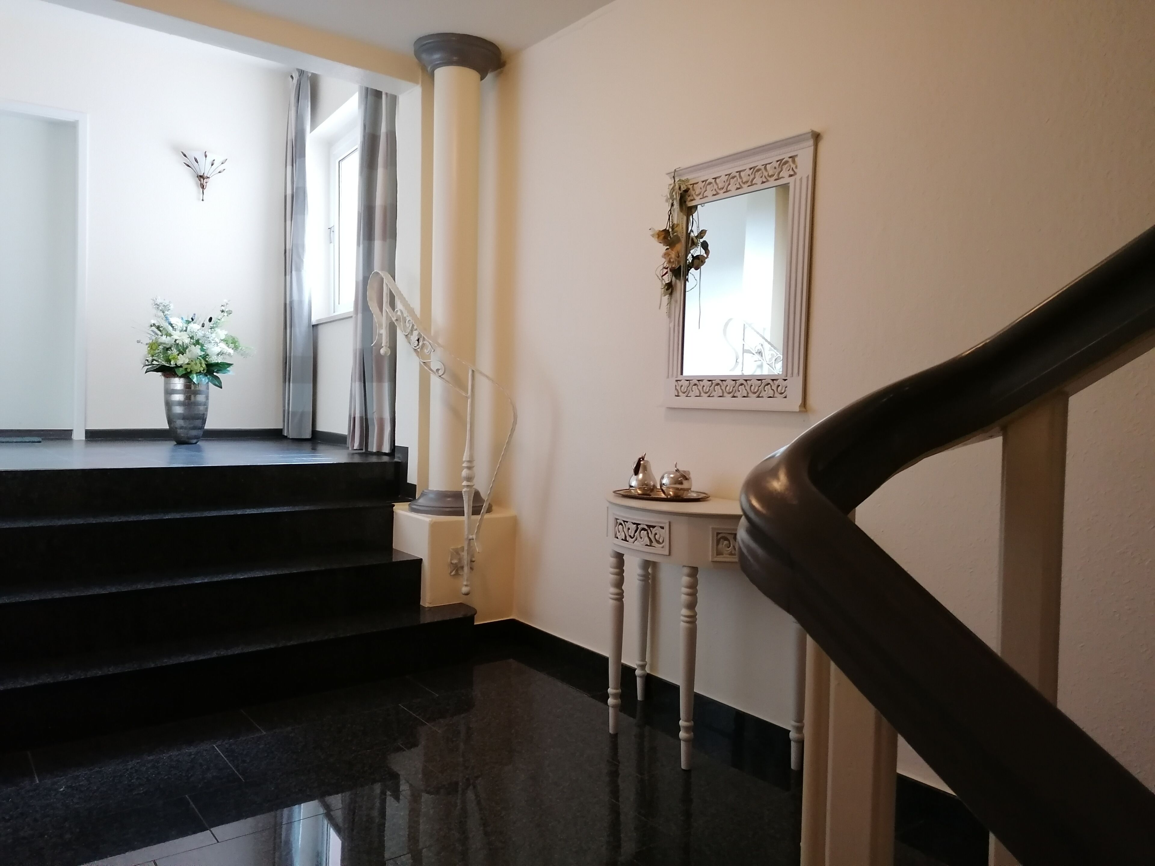 Eine glatte schwarze Treppe führt zu einer Empore mit einer weißen Säule, einem kleinen Tisch mit Dekoration und einem Spiegel an der Wand.