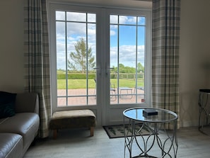 Ein Zimmer mit einer Couch, einem Hocker, einem Glastisch und Blick durch eine Tür auf eine Terrasse im Freien mit Rasen und Baum.