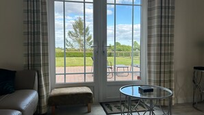 Ein Zimmer mit einer Couch, einem Hocker, einem Glastisch und Blick durch eine Tür auf eine Terrasse im Freien mit Rasen und Baum.