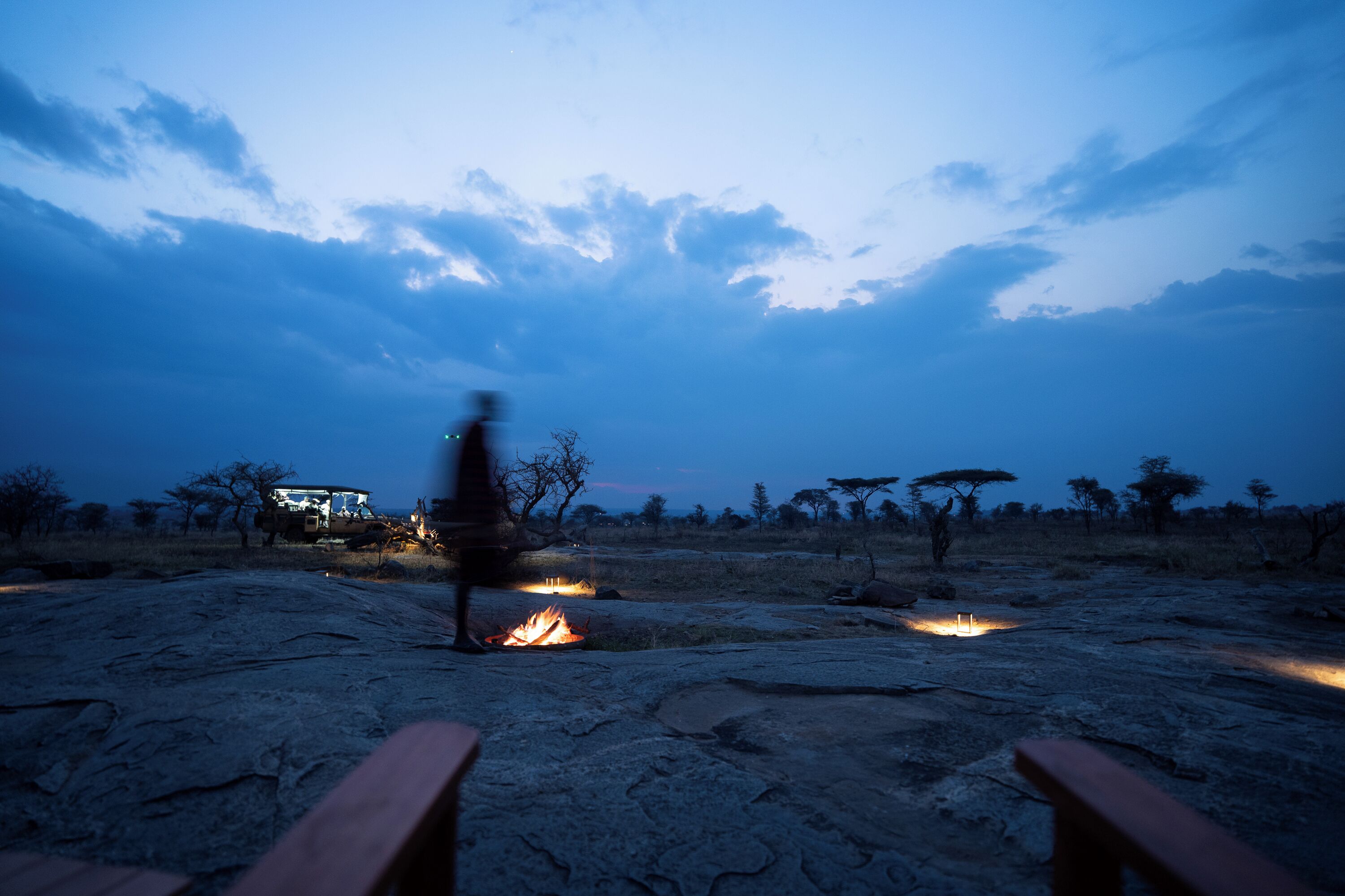 A campsite with a fire, a vehicle, and a person walking.