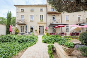 A building with a central entrance, outdoor seating with umbrellas, and a garden with hedges and potted plants. - Boutique Hôtel La Villa Montesquieu (Laudun-l'Ardoise)