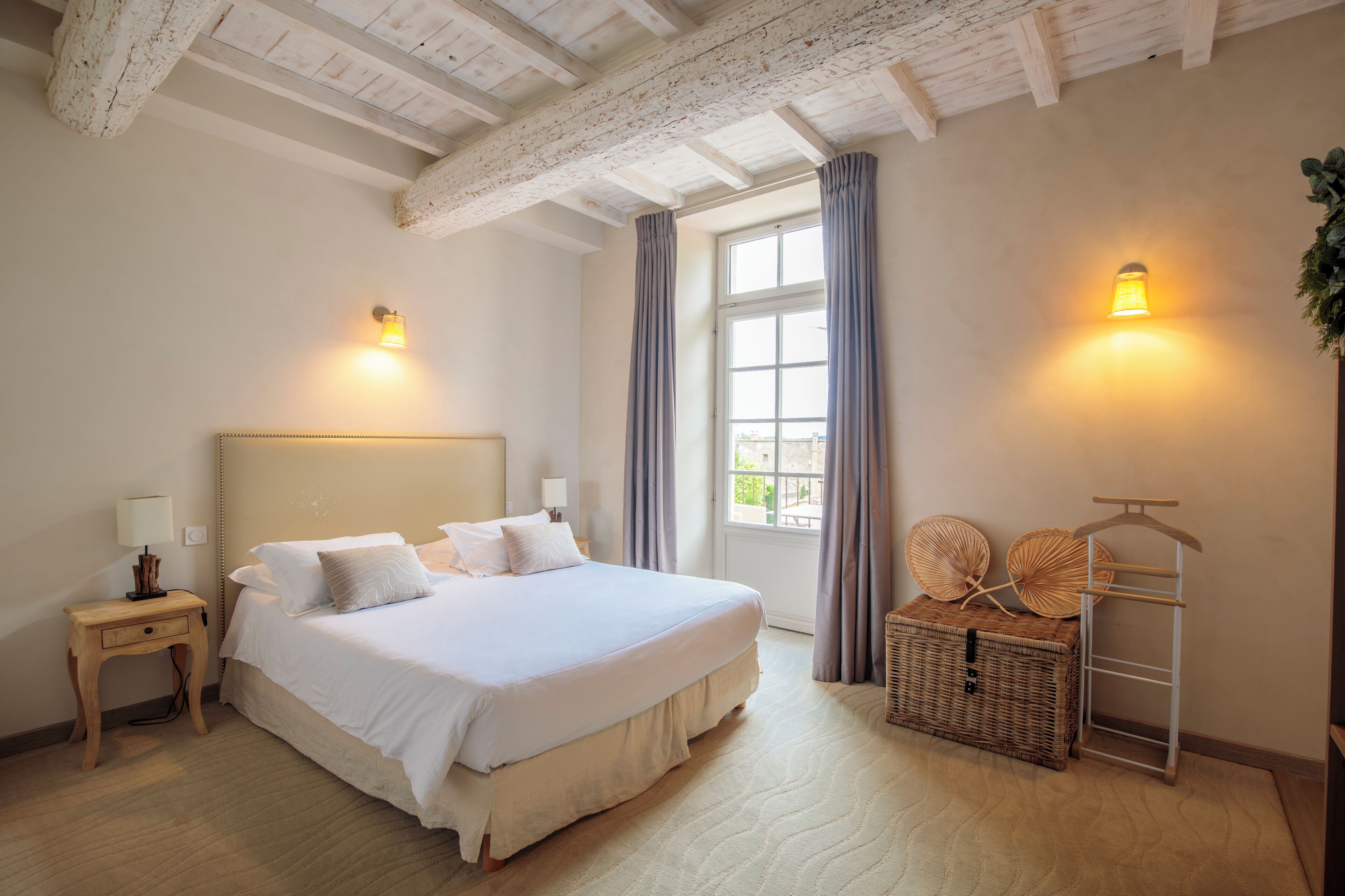 A bedroom with a large bed, a bedside table, a window with curtains, and a wall-mounted light fixture.