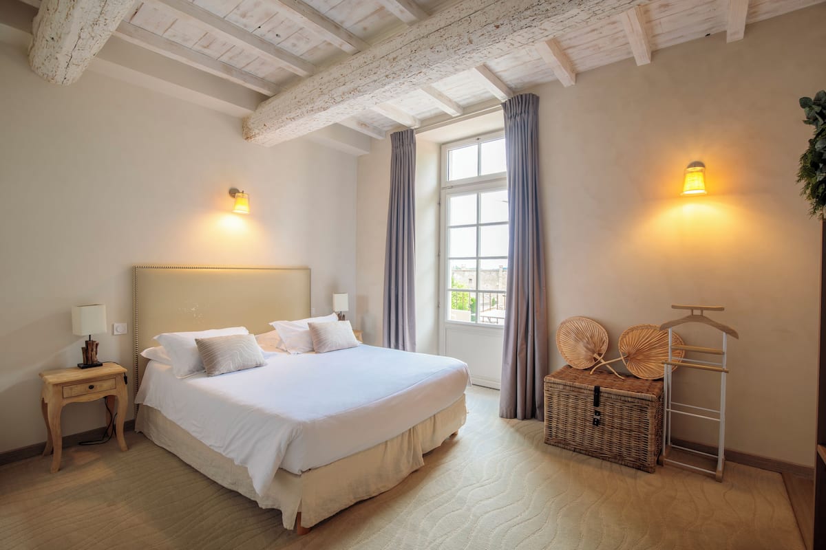 Un dormitorio con una cama grande, una mesita de noche, una ventana con cortinas y una lámpara fijada a la pared.