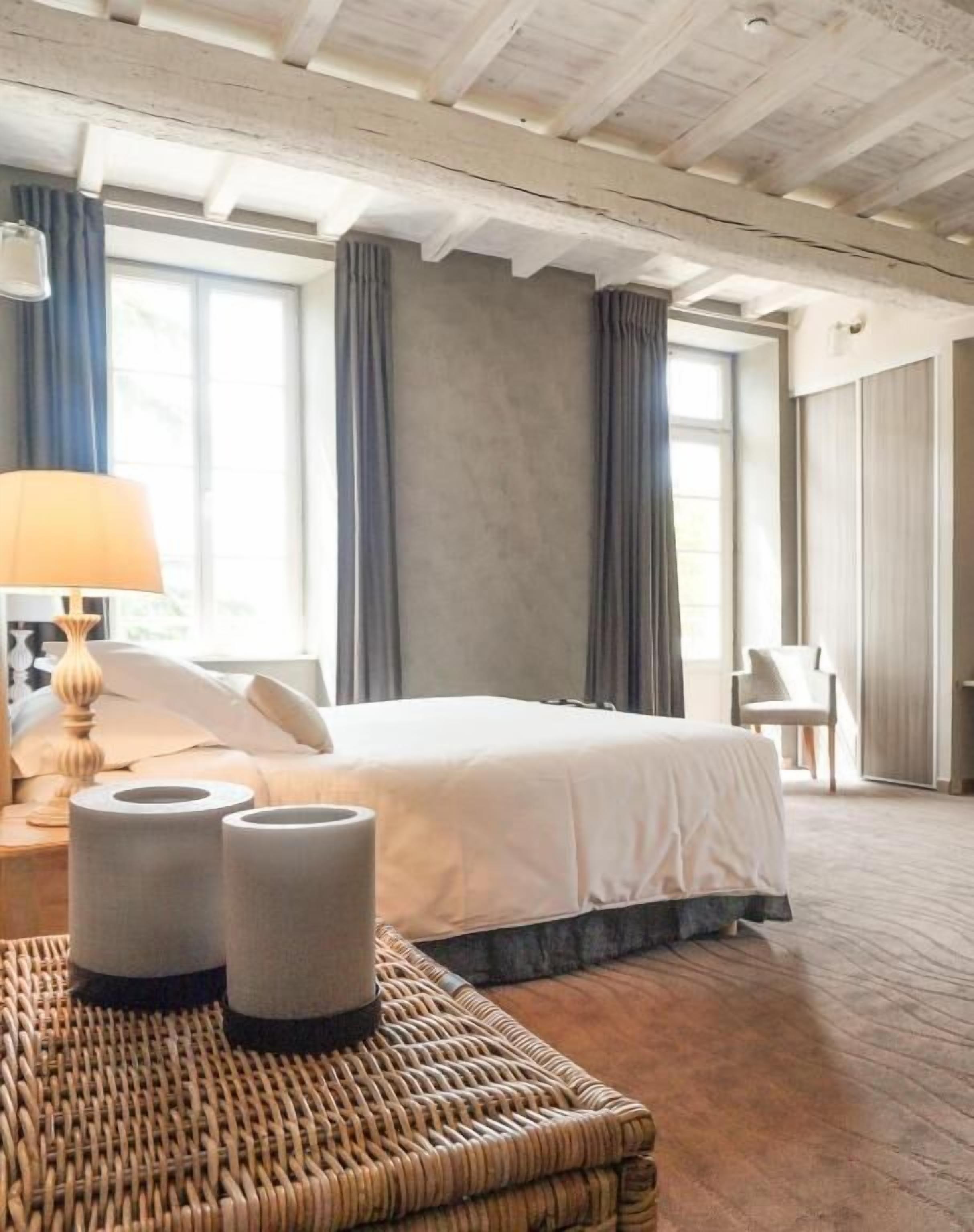 A hotel room with a large bed, a wicker side table, a lamp, and a view of the outdoors through the window.