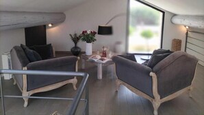A modern loft-style room with a high ceiling, exposed wooden beams, a large window, and two grey upholstered armchairs. - Boutique Hôtel La Villa Montesquieu (Laudun)
