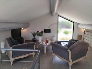 Una habitación moderna estilo loft con techo alto, vigas de madera a la vista, un ventanal grande y dos sillones grises tapizados.