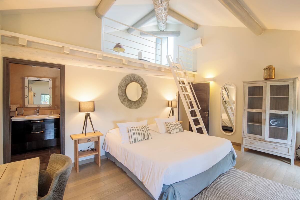 Un dormitorio con techo estilo loft, una cama con ropa de cama blanca, una mesita de noche, un espejo, un armario y un pequeño escritorio con silla.