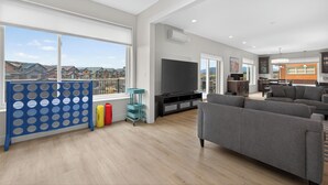 Een ruime woonkamer met een grote televisie, grijze banken en uitzicht op huizen.