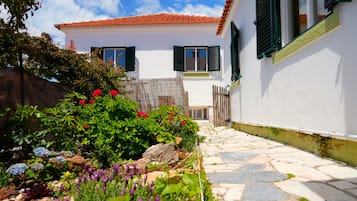 Um edifício branco com portadas verdes, um caminho de pedra e um jardim com diversas plantas e flores.
