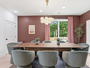 Ein Esszimmer mit Holztisch, sechs Polsterstühlen, einer Hängelampe, einem gerahmten Bild an der Wand und Blick ins Freie durch ein Fenster.
