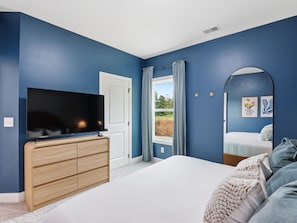 Un dormitorio con una televisión sobre un aparador de madera, una cama con colchón blanco y vistas a la vegetación a través de una ventana.