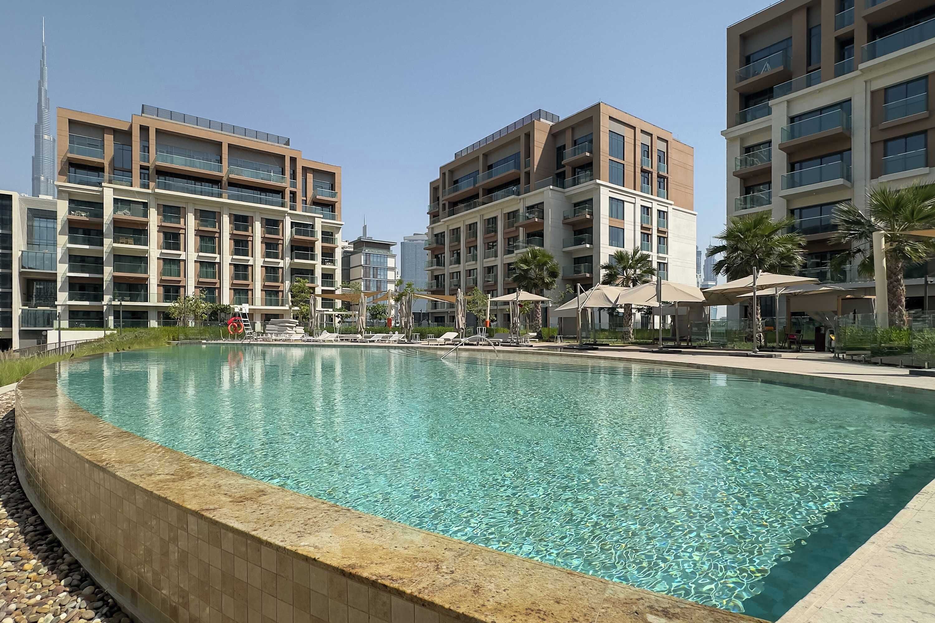Una piscina rodeada de edificios modernos con balcones, cielo despejado y palmeras.
