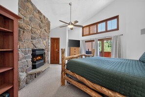A bedroom with a stone fireplace, a bed with a green quilt, a ceiling fan, and a view of the outdoors through a window.