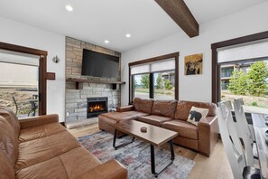 A modern living room with a stone fireplace, leather sofas, a TV, and large windows.