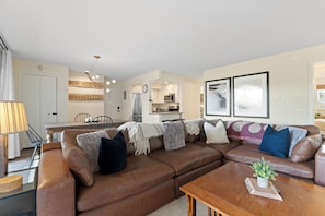 A living room with a brown leather sofa, a wooden coffee table, and a kitchen in the background. - The Colony Unit #102 Ground floor, renovated, private patio, near outdoor trails (Fraser)