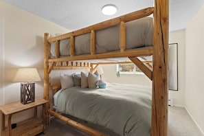 A bunk bed with a wooden frame, a bedside table with a lamp, and a window with a view. - The Colony Unit #102 Ground floor, renovated, private patio, near outdoor trails (Fraser)