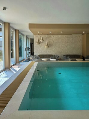 A modern indoor pool with clear glass walls, a stone-textured wall, and a seating area with chairs and a sofa.