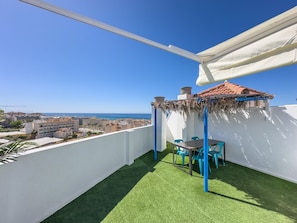 Un balcón con césped artificial, una mesa con sillas, un dosel blanco y vistas al mar y edificios.