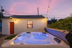 Un jacuzzi con cubierta de madera, un edificio pequeño con puerta roja y luces colgantes.