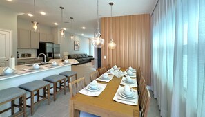 Eine moderne Küche mit einer zentralen Kochinsel, ein Essbereich mit einem langen Tisch für sechs Personen sowie eine Wohnbereich mit Sofa und Fernseher.