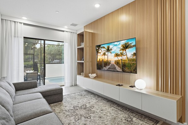 A modern living room with a flat-screen TV mounted on a wooden wall, a grey sofa, and a patterned rug. - Luxury 5BR Villa w/ 4 Private Suites | Heart of Orlando (Orlando)