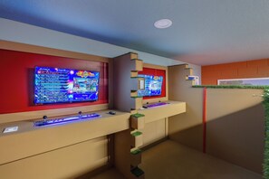 Une pièce avec deux consoles de jeux fixées sur un mur, une petite étagère avec une télécommande, et un mur d’accent de couleur rouge.
