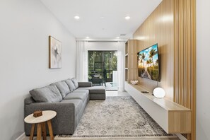 Ein modernes Wohnzimmer mit einem grauen Ecksofa, einem kleinen runden Tisch, einem gemusterten Teppich, einem Flachbildfernseher, der an einer Holzwand befestigt ist, und Blick durch eine Glastür auf einen Außenbereich.