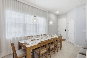 Une salle à manger avec une table en bois dressée pour six personnes, deux lampes suspendues, une porte blanche et une fenêtre ornée de rideaux légers.