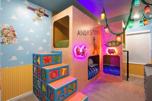 Une salle de jeux pour enfants avec des blocs d’alphabet colorés, un lit avec un jouet en forme de dinosaure et un mur orné d’une enseigne « Les jouets d’Andy ».