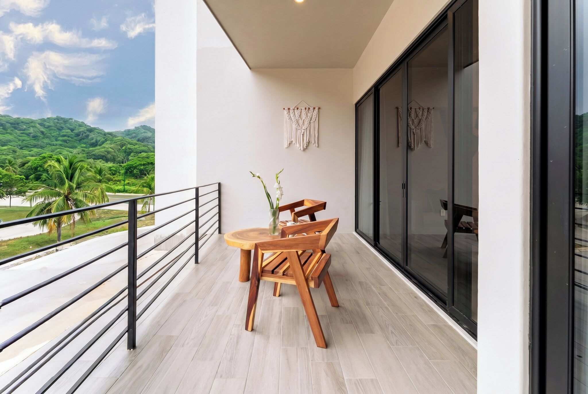 A balcony with a wooden table and chairs, a macrame wall hanging, and a view of a green landscape.