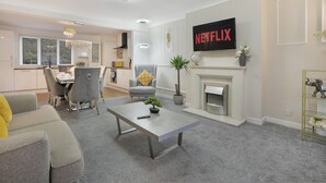 Een moderne woonkamer met een open haard, een televisie waarop 'NETFLIX' staat, en een eethoek met een kroonluchter.