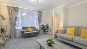 Een woonkamer met een grijze bank, een bijzettafel met een bonsai, een grijze fauteuil, een lamp en een raam met gordijnen.