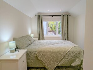 Ein Schlafzimmer mit einem Bett, zwei Nachttischlampen, einem Fenster mit Vorhängen und Blick auf Grünfläche.