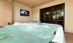 Ein moderner Wellnessraum mit Whirlpool, einem an der Wand befestigten Fernseher und einer großen Schiebetür aus Glas.