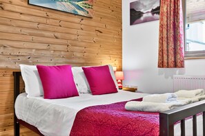Habitación de hotel con pared de madera, cama con ropa de cama blanca y rosa, una manta roja y una ventana con cortinas.