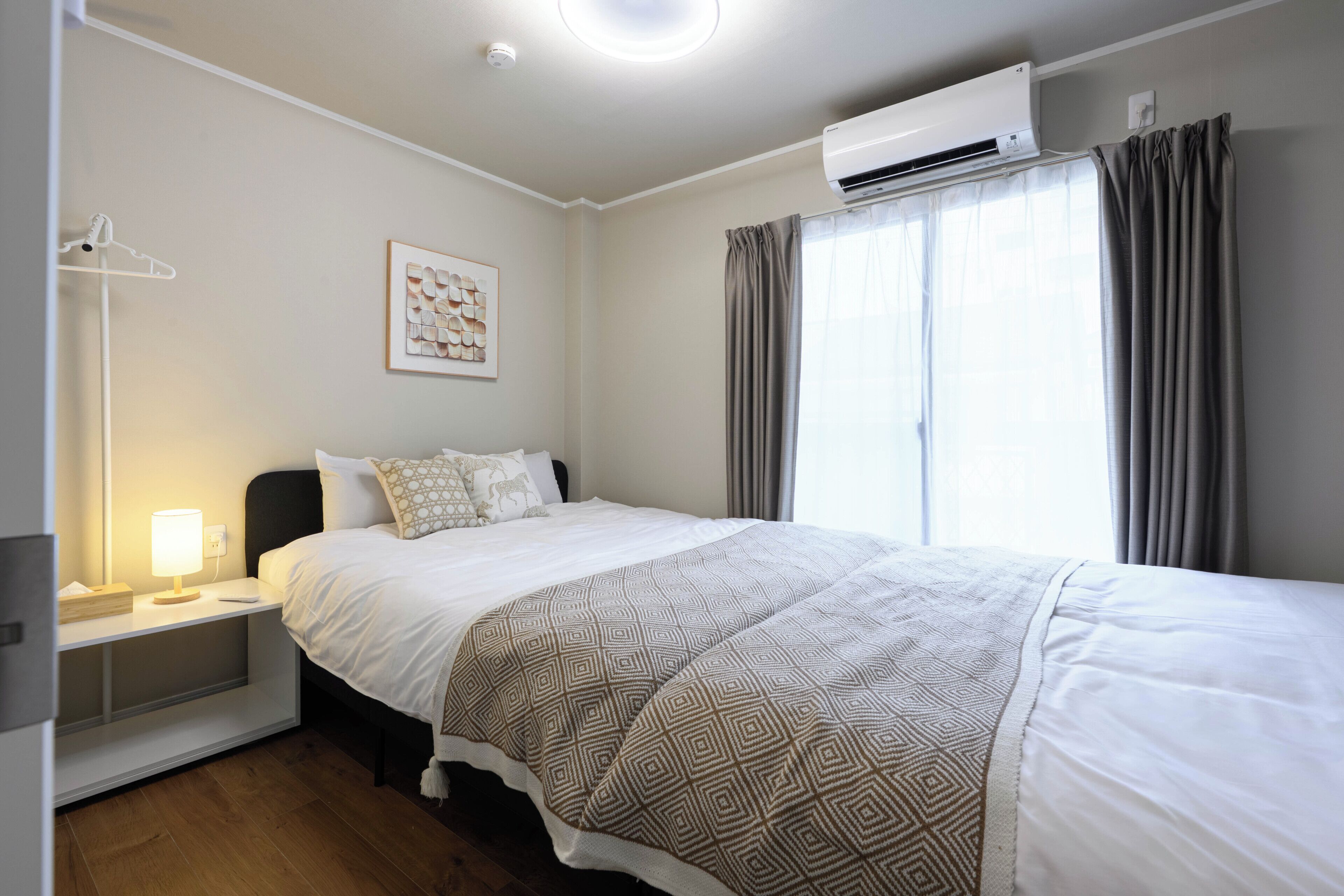 A hotel room with a bed, bedside table, lamp, air conditioning unit, and a window with curtains.