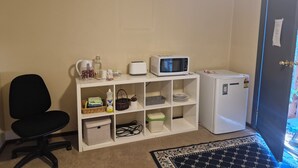 Una habitación con aire acondicionado blanco, un microondas blanco, un refrigerador blanco, una silla de oficina negra y una estantería blanca con varios objetos.