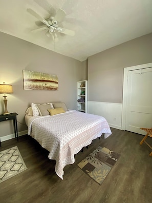 A bedroom with a bed, a ceiling fan, a lamp, a painting, a bookshelf, and a door.