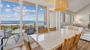 Een ruim balkon met uitzicht op de oceaan, een eethoek met tafel en stoelen, en een modern interieur met een groot raam en een hanglamp.