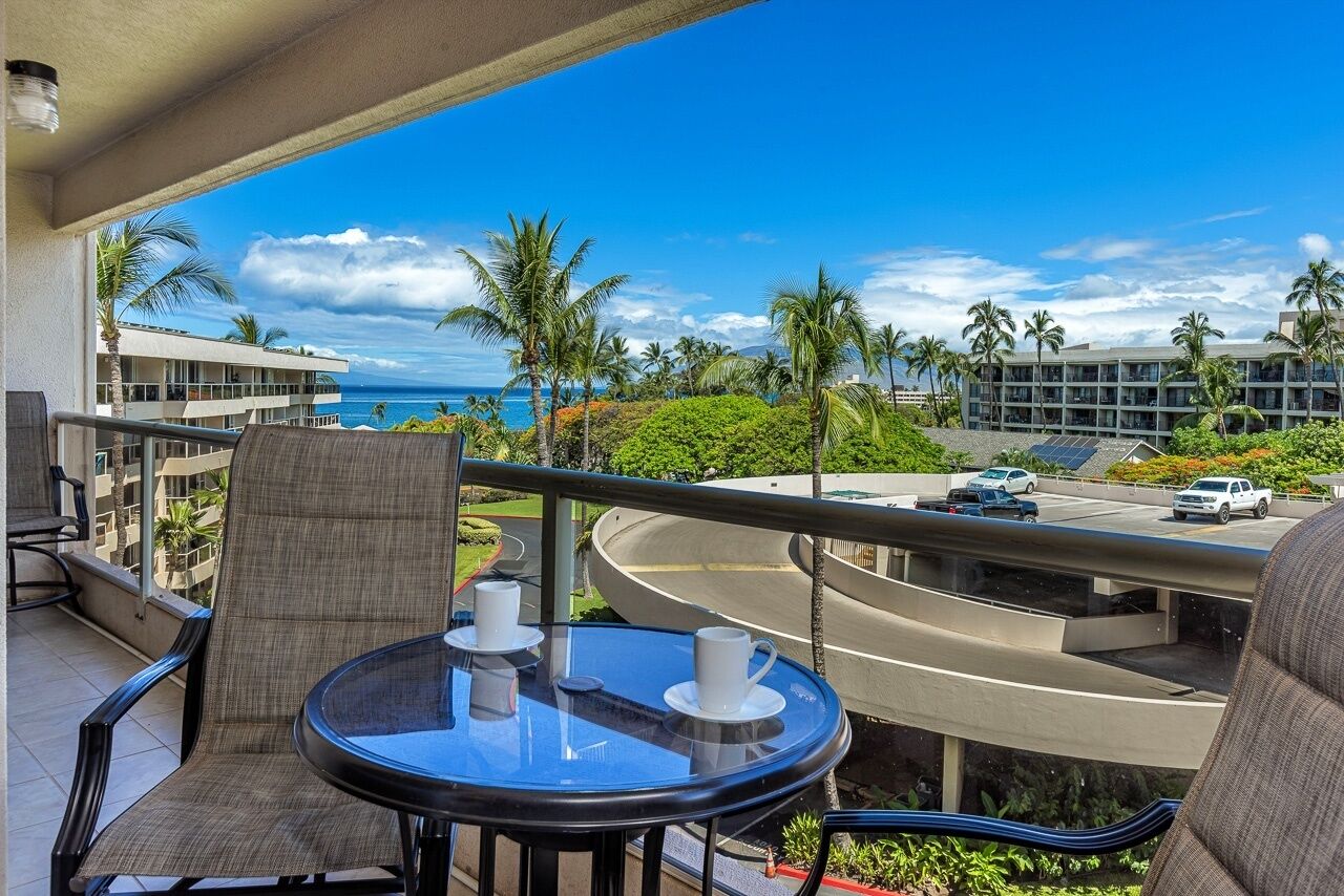 A balcony with a table, chairs, and a view of the ocean and palm trees.
