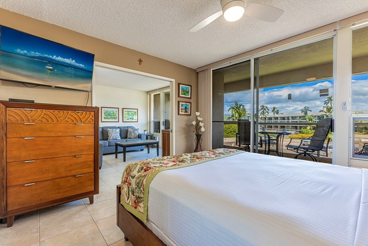 A hotel room with a bed, a dresser, a TV, a balcony with a view of palm trees, and a dining area.