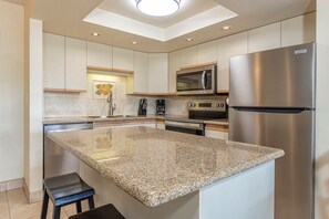 A modern kitchen with a central island, stainless steel appliances, and a recessed ceiling light. - Maui Banyan H406 1 Bedroom Condo (Kihei)